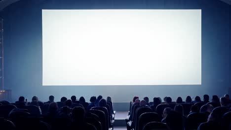 audience members sit in theater seats, focused on a bright white screen in a dark cinema environment