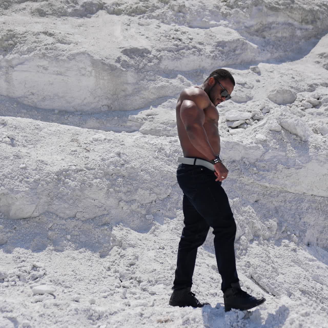 Vista di una telecamera in movimento di un uomo americano allegro a torso nudo che cammina sulla spiaggia sabbiosa. Uomo muscoloso attraente e affascinante con concetto di torso nudo