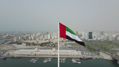 uae flag: drone camera circling around the flag of united arab emirates waving in the air at sharjah's flag island, sharjah city in the background