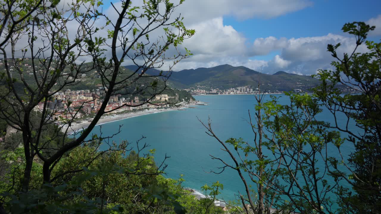 Noli, Liguria: rapido zoom in da rami in primo piano a un'ampia baia turchese e città di mare sullo sfondo di montagne; dinamico panorama costiero in una giornata luminosa