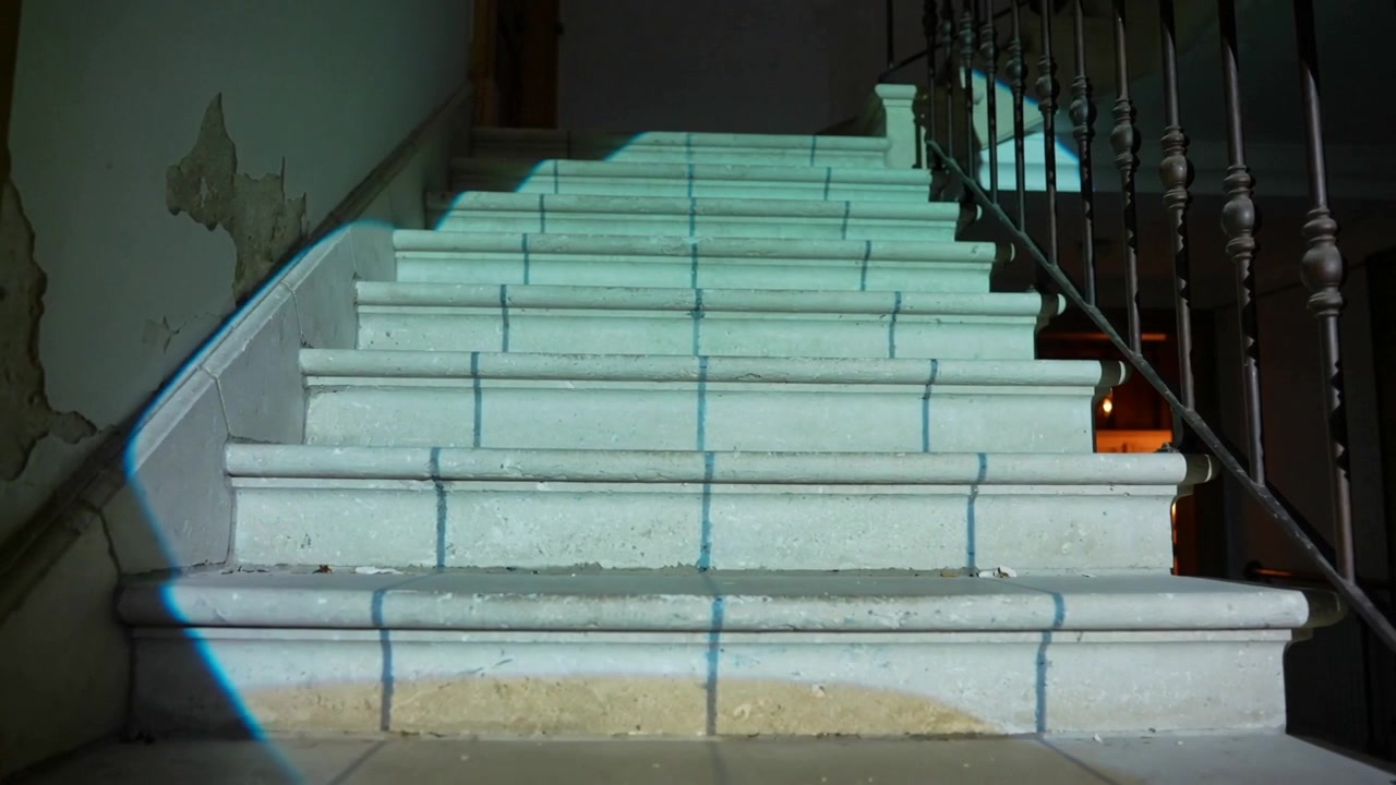 Una proiezione blu appare e sale lungo le scale piastrellate sul pianerottolo, rivelando una balaustra e un caldo bagliore.
