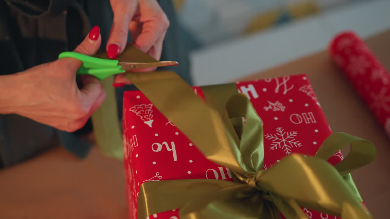 Manos cortando una cinta verde en un regalo, primer plano de tijeras recortando los extremos de la cinta. Envoltorio festivo rojo con motivos blancos, un acabado práctico del regalo y una preparación festiva eficiente en un espacio de trabajo moderno.