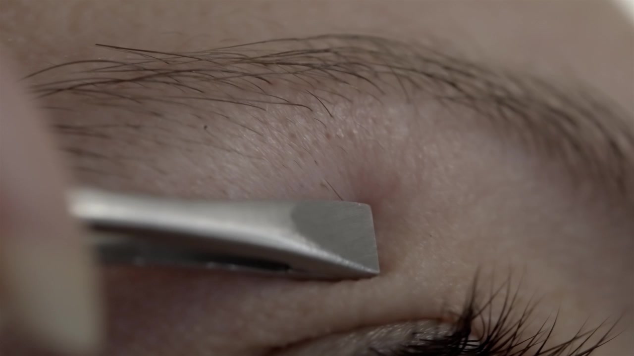 Fokuseret billede af en kvinde, der omhyggeligt former sit øjenbryn ved hjælp af en metalpincet.