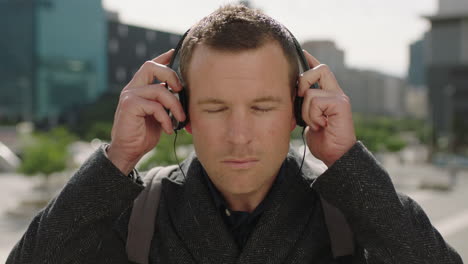 portrait of attractive young caucasian businessman looking serious at camera puts on headphones listening to music in sunny urban city