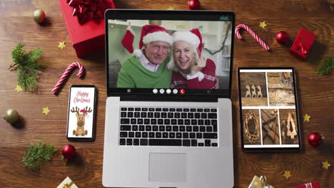 Caucasian-senior-couple-waving-on-video-call-on-laptop,-with-smartphone,-tablet-and-decorations