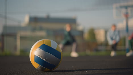 close-up of volleyball on ground with blurred background showing athletes exercising and stretching outdoors, a building is visible in the distance with vibrant sunlight