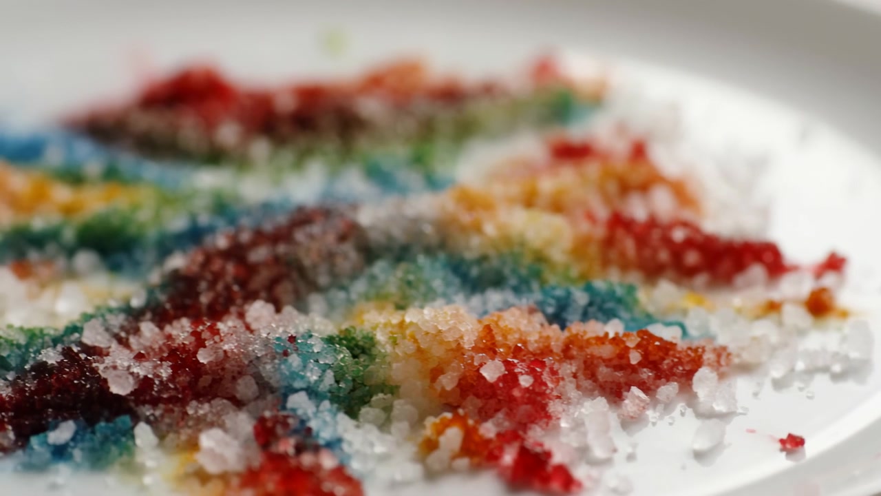 Cristais de açúcar grosso coloridos dispostos em fileiras diagonais em um prato de cerâmica branco, molhando e dissolvendo lentamente enquanto os pigmentos se espalham entre os grânulos translúcidos, criando uma textura brilhante e um efeito abstrato.