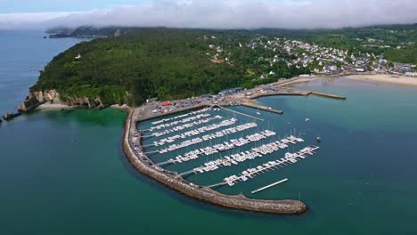 drone pulling back over morgat marina in crozon, showing the harbor, breakwater, coastline, vegetation and distant houses - brittany france