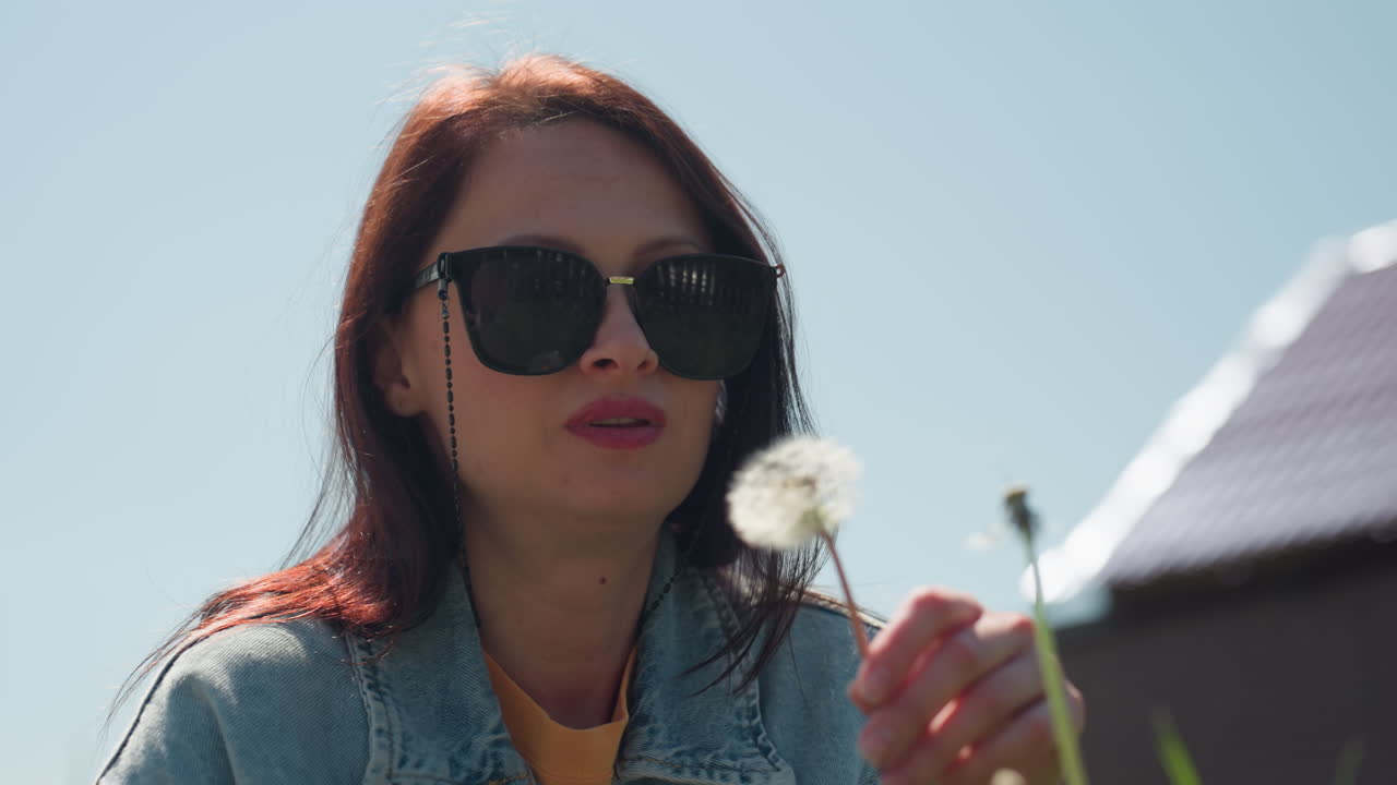 Young woman wearing sunglasses and denim jacket gently blows dandelion in sunlight, holding stem close to face with lips pursed, set against clear blue sky and roof background