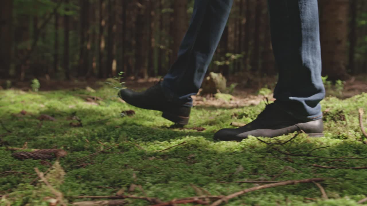 A low-angle close-up shot follows a man's feet in black boots as he walks across a lush, green carpet of moss on the forest floor. Patches of sunlight create a peaceful and serene atmosphere.