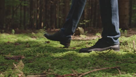 a low-angle close-up shot follows a man's feet in black boots as he walks across a lush, green carpet of moss on the forest floor. patches of sunlight create a peaceful and serene atmosphere.