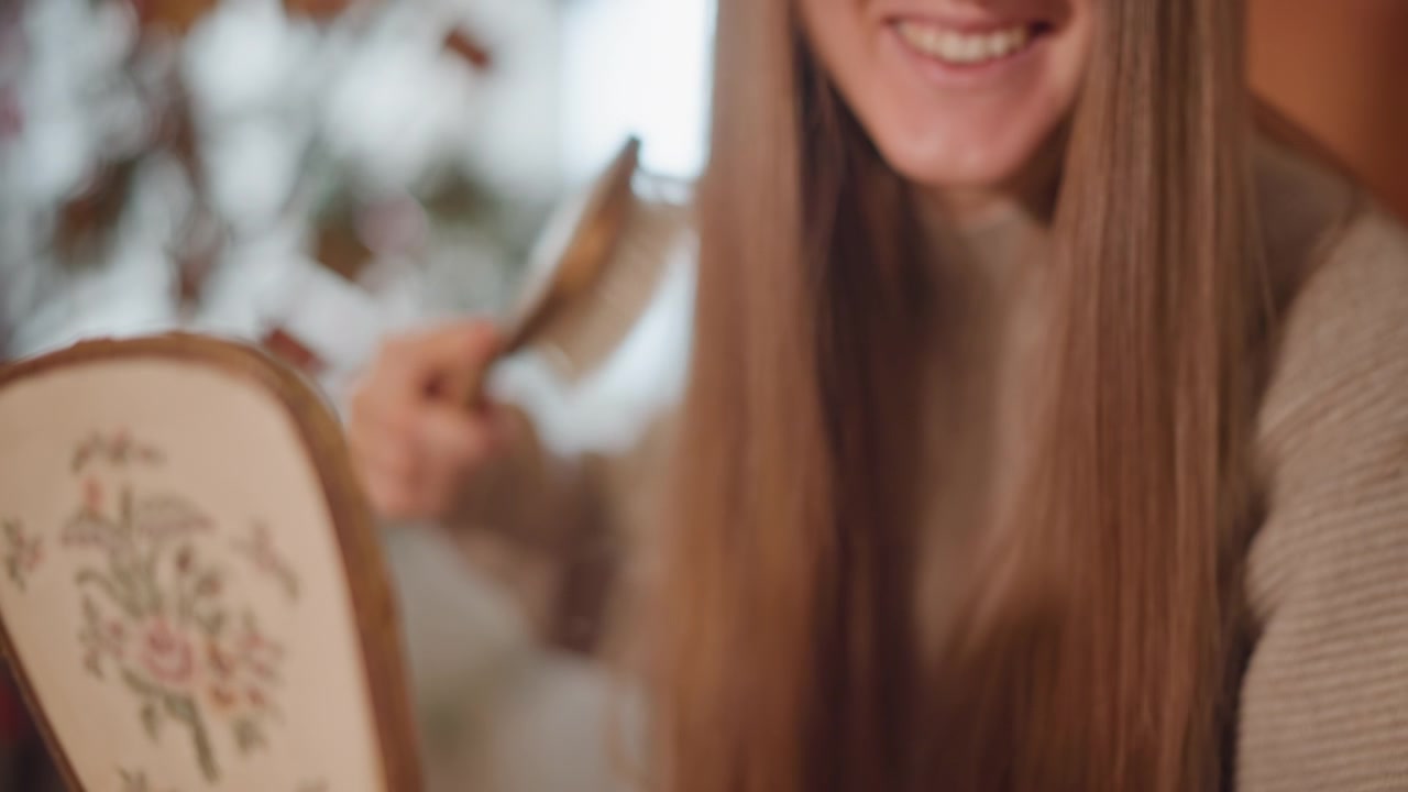 Reflejo de una dama retro. Mujer con cabello largo sonriendo. Mujer sonriente con espejo vintage cerca de plantas otoñales. Escena nostálgica de una mujer cepillándose el cabello largo junto a una ventana soleada con detalles bordados
