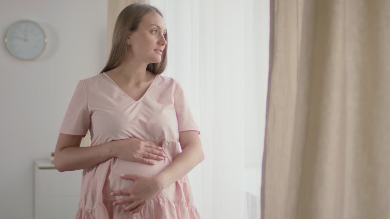 mujer en la habitación de su bebé tocándose la barriga y mirando por la ventana