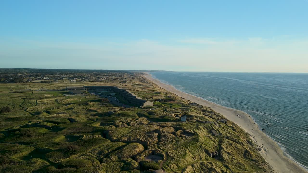 En drone flyver i et filmisk shot langs Nordsøkysten mod Fyrklit Hotel og viser de store sandklitter, kystvegetationen og de gyldne solnedgangsrefleksioner på vandet i Nordjylland