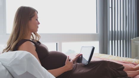 Mujer-Embarazada-Relajándose-En-La-Cama-En-Casa-Viendo-Una-Tableta-Digital-Sosteniendo-El-Estómago