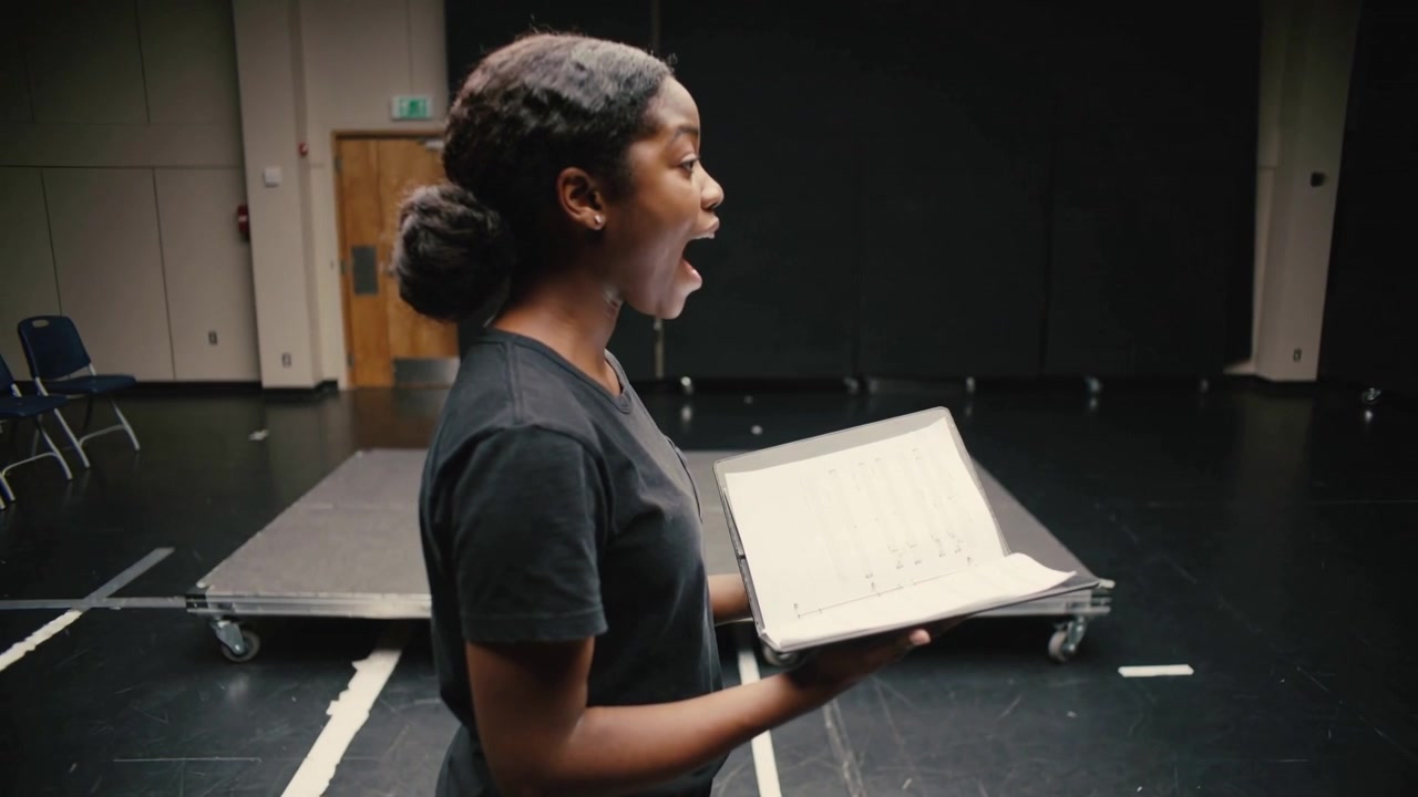 Un artiste en T-shirt noir prend une inspiration et projette sa voix en lisant un classeur, dans une salle de répétition, répétant sa performance.