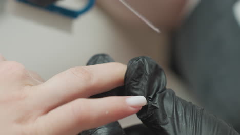 closeup of nail technician wearing black gloves filing client fingernail while gently brushing away nail dust scene highlights careful attention to hygiene, detail, and client nail shaping in salon