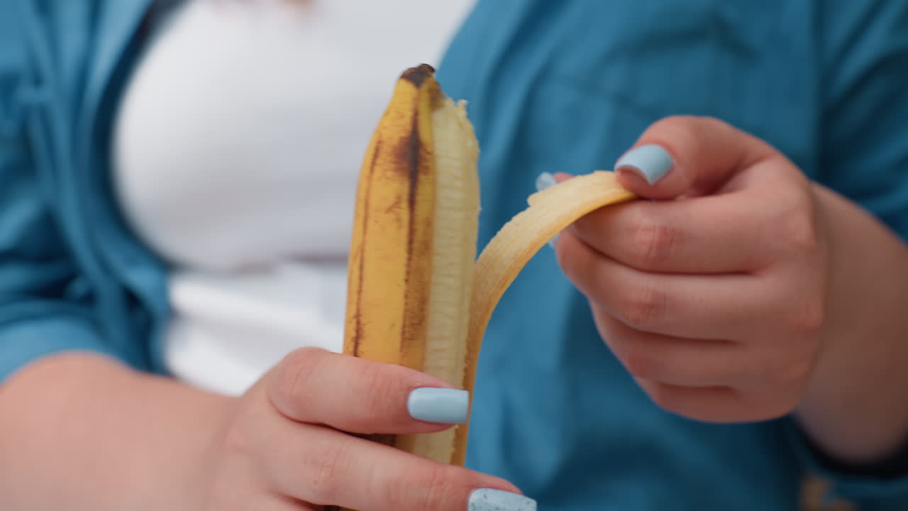 Primer plano de una mujer pelando la piel de un plátano, revelando la fruta fresca; la luz natural enfatiza la superficie amarilla lisa y la textura suave, movimiento delicado de las manos con esmalte de uñas azul claro