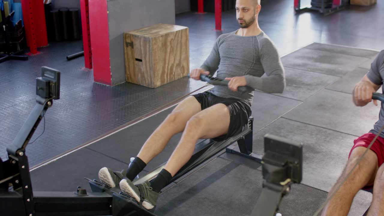 Diverse male gym partners adjusting straps grabbing water bottles and rowing on rowers for sparring