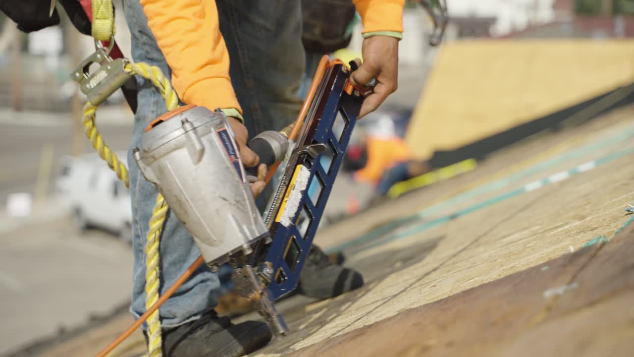 trabajador usando una pistola de clavos para asegurar nuevas tablas de madera contrachapada en un techo residencial durante la renovación.
