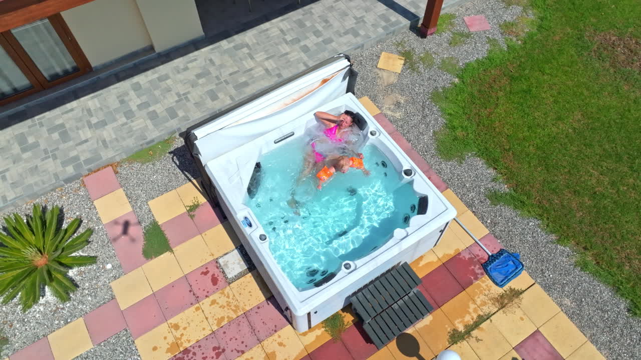 High angle drone shot circling a mom and son relaxing in a jacuzzi, sunny day