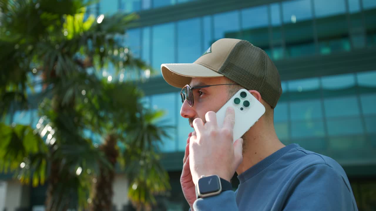 hombre joven con gorra de béisbol y gafas de sol tomando una llamada telefónica fuera del edificio de oficinas de vidrio