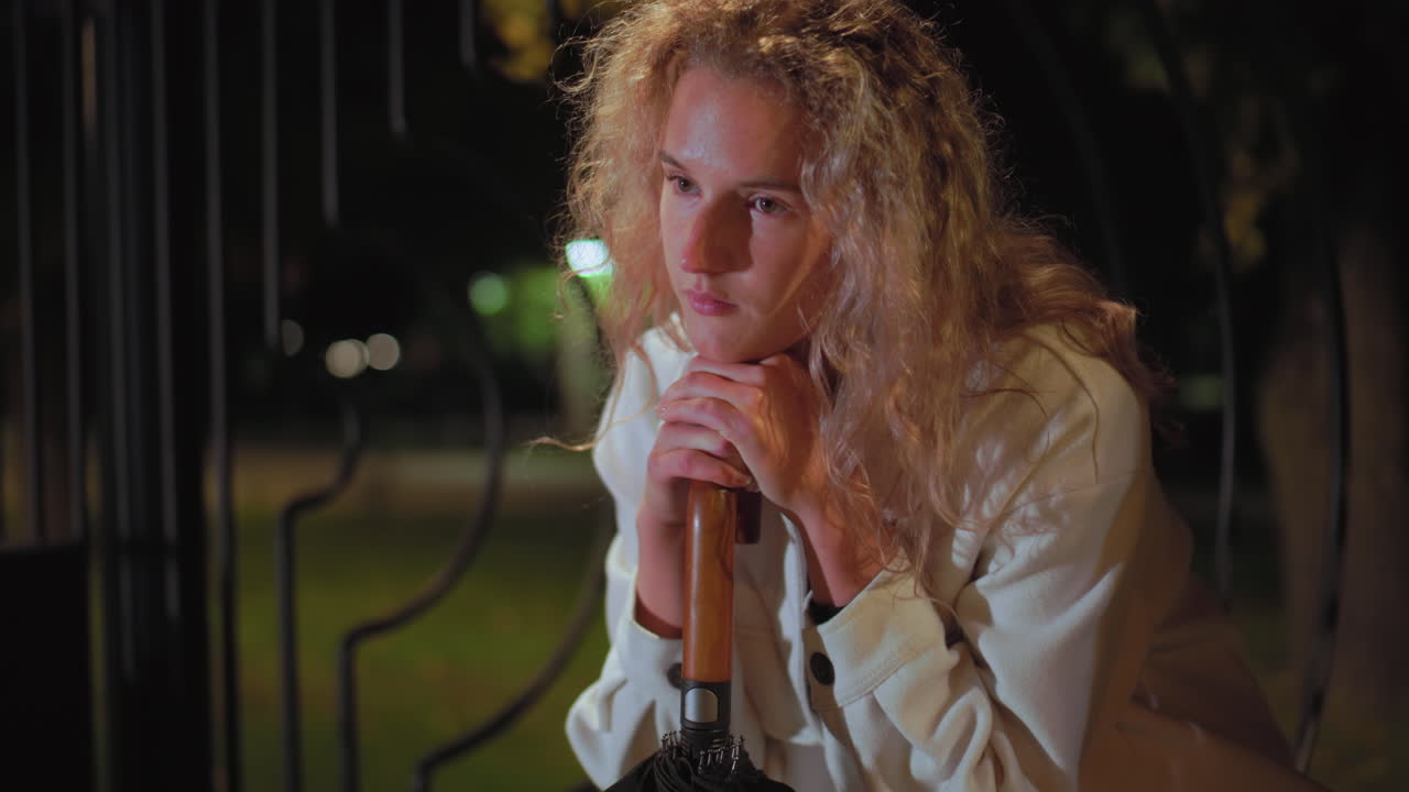 Young girl with curly hair wearing white coat sits quietly at night, resting head on umbrella handle, looking exhausted and peaceful under soft glow of street lights with calm park background
