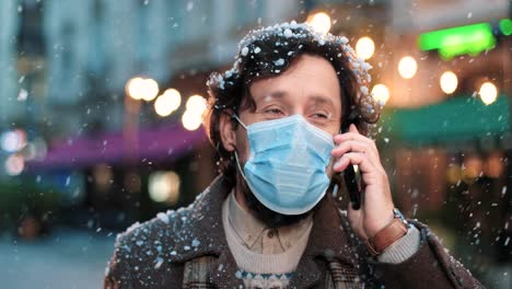 close-up view of caucasian man wearing a face mask and talking on smartphone on the street while it‚äôs snowing in christmas