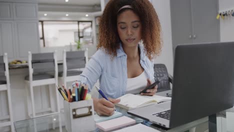 busy biracial woman using smartphone and laptop, writing notes at home, in slow motion