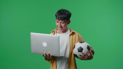 niño asiático con una pelota viendo un partido de fútbol en una computadora portátil en el estudio de pantalla verde