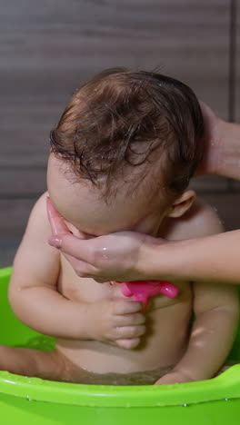 caring mom's hands washing her son sitting in the washbowl. adorable baby boy playing with a rubber toy while washing. vertical video