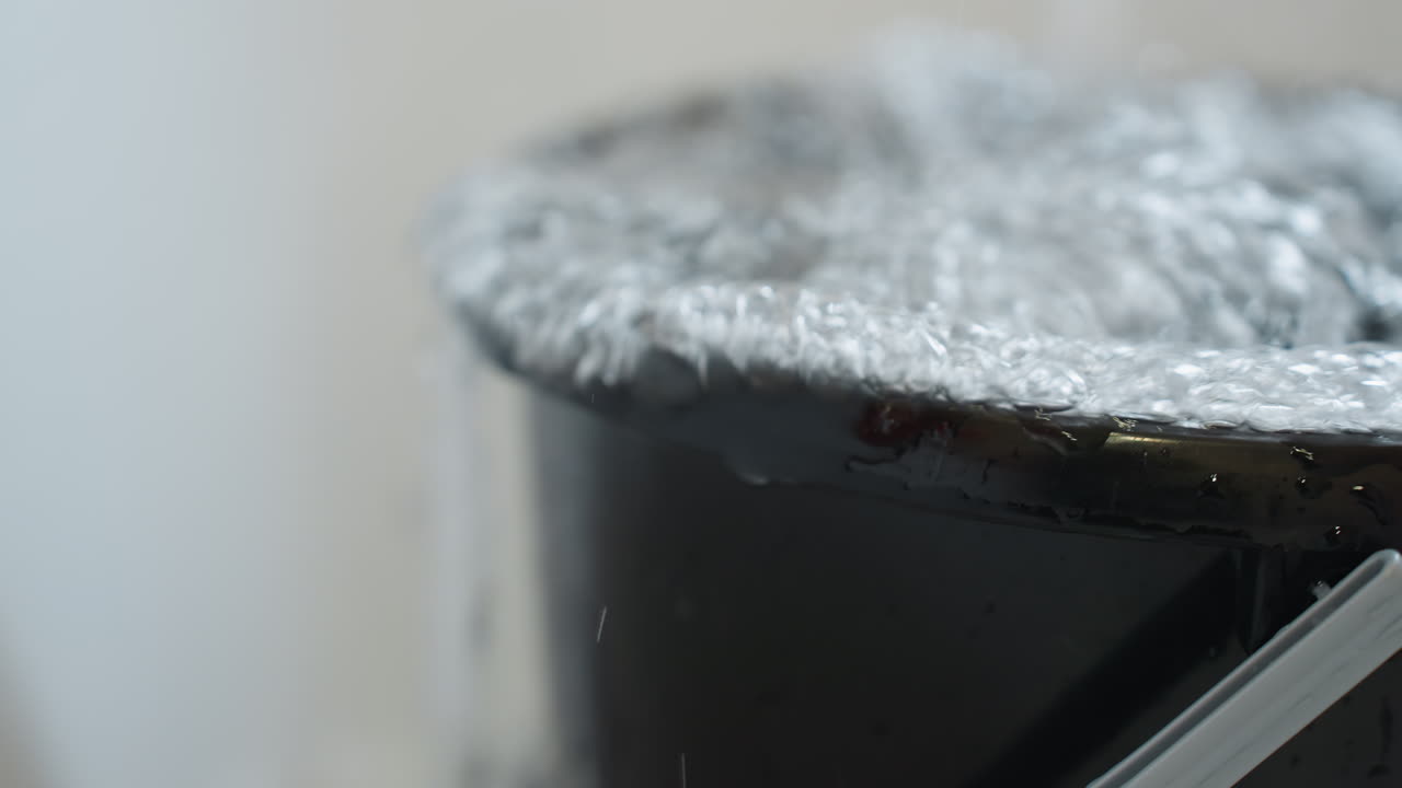 Close up of black bucket completely filled with water as liquid pours out from top edges, surface forming bubbles and ripples, concept of overflow, waste, household usage and cleaning