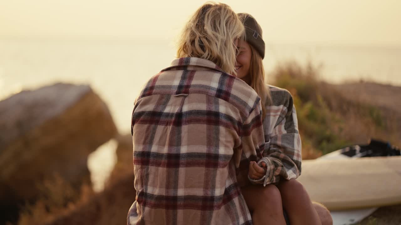A blonde girl in a black hat and a checkered shirt sits on the lap of her blonde boyfriend in a checkered shirt on the seashore Sunny morning at sunrise