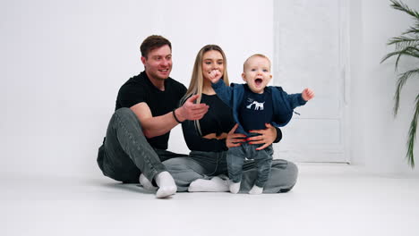 caucasian parents sit on the floor holding a baby boy. little kid is cheering and holding hands up. white backdrop.