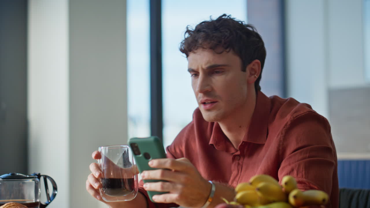 Disappointed guy looking cellphone receiving bad news at home kitchen closeup