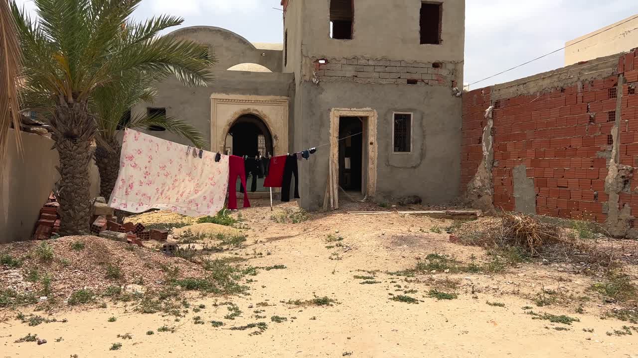 Klær og plagg henger og tørker på utendørs klessnor i en tunisisk landsby med et forfallent eller uferdig hus i bakgrunnen