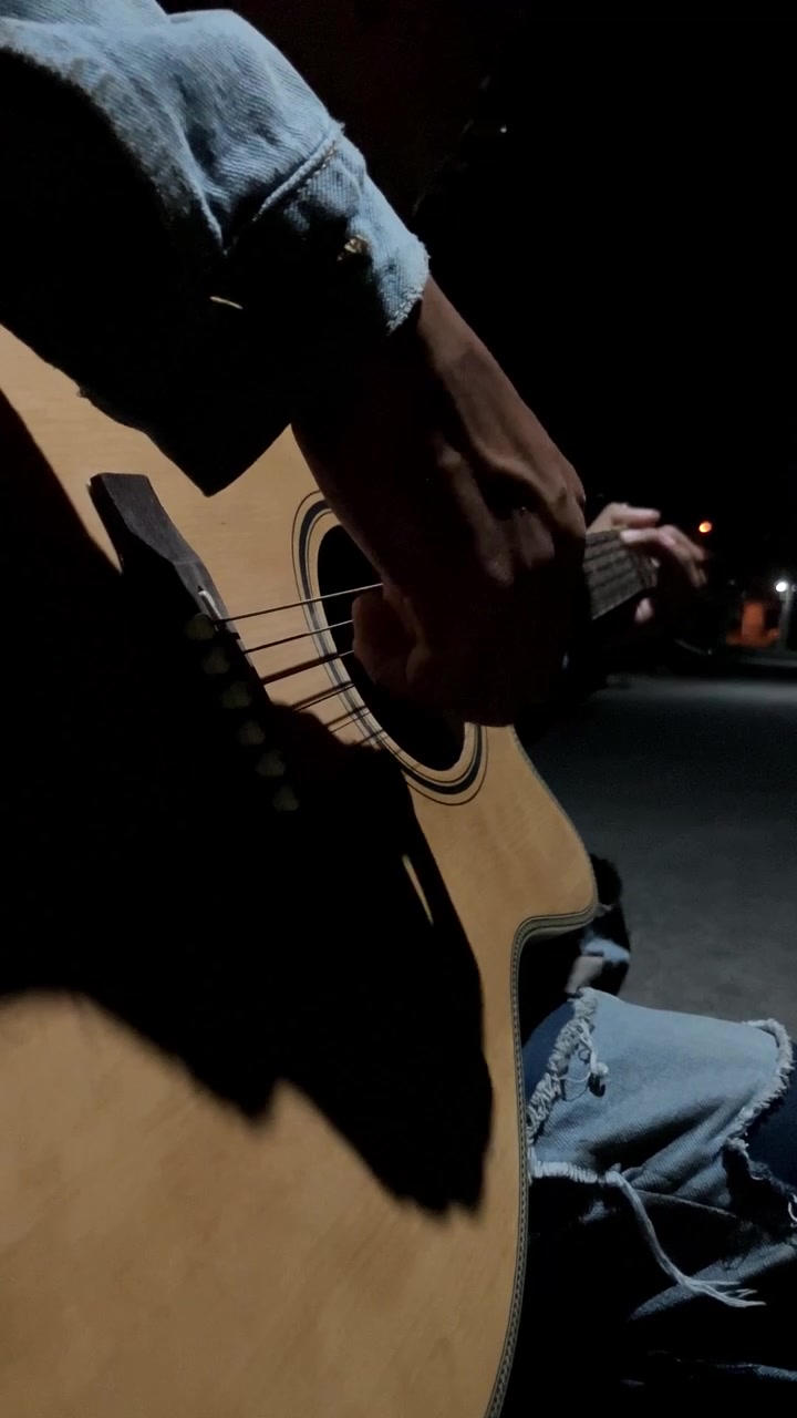 Vertikalt 4K nærbillede med fokus på en mands højre hånd, der spiller på en akustisk guitar om natten. Mørk, stemningsfuld low key belysning skaber en intim, æstetisk musikalsk performance b-roll.