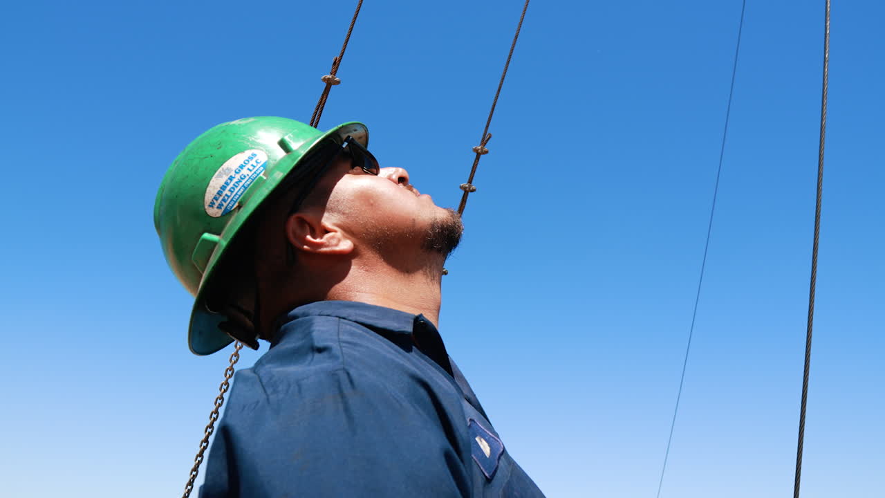Bearded man wearing sunglasses and protective helmet looks up and gestures with his hand. Portrait of a worker at the site for drilling oil. Low angle view.