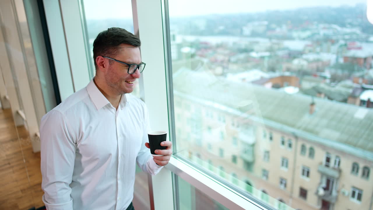 Break from work in the office. Smiling man drinking coffee standing at the window looking at cityscape.