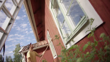 man brushes off peeling falu red paint from exterior wall, house maintenance