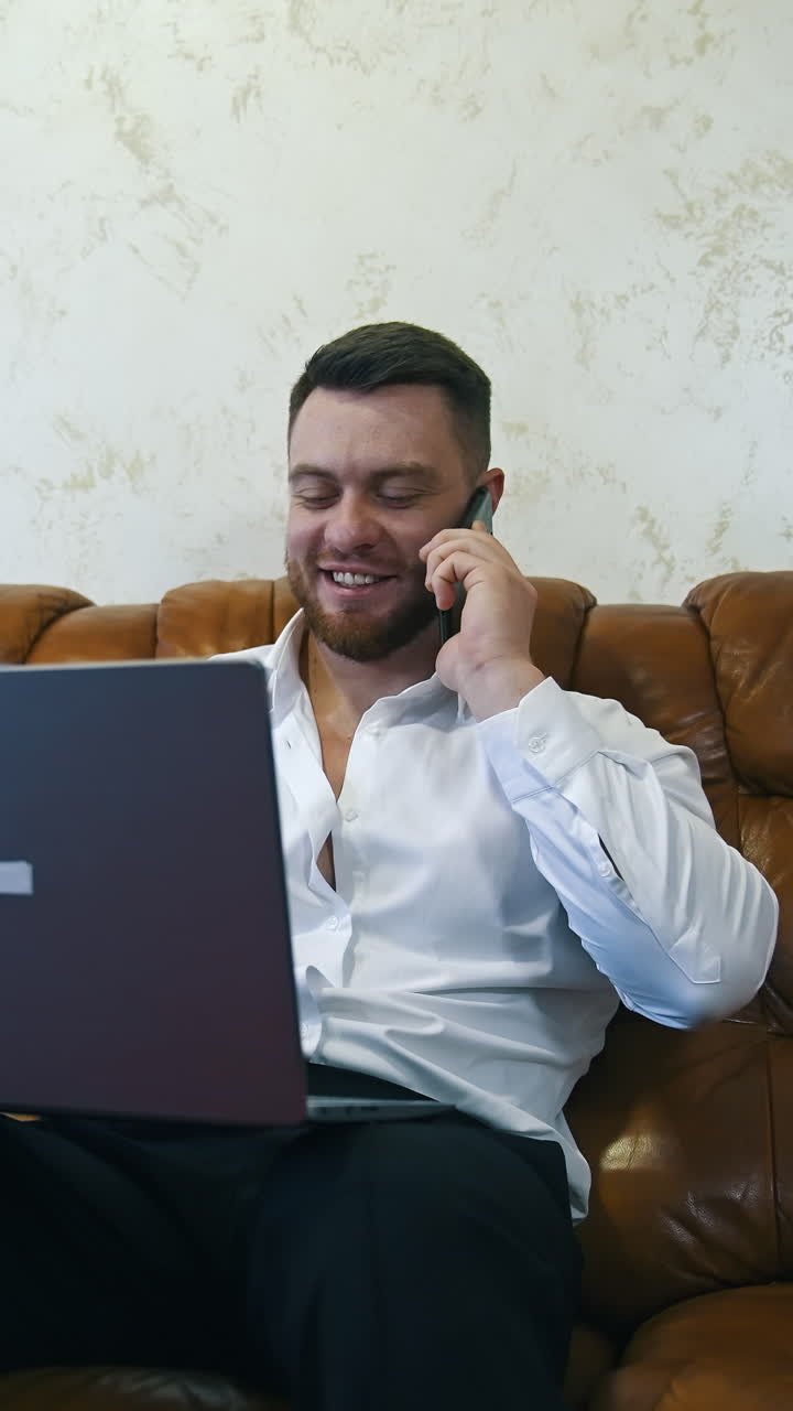 Relaxed businessman is working in a hotel room on laptop. Handsome man with laptop on knees speaks on the mobile phone. Vertical video