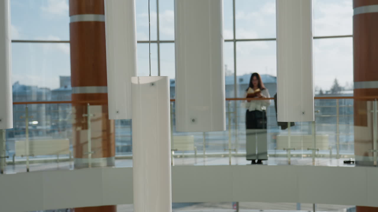 Decorative vertical ceiling lights dominate foreground as young girl stands reading near glass railing in bright indoor gallery with large windows and urban buildings in soft background focus