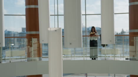 decorative vertical ceiling lights dominate foreground as young girl stands reading near glass railing in bright indoor gallery with large windows and urban buildings in soft background focus