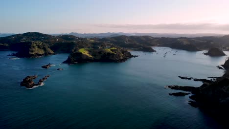 Scenic-View-Of-Calm-Blue-Sea-And-Green-Rocky-Hills-At-The-North-Coast-Of-New-Zealand---Tutukaka-On-A-Golden-Hour-Sunset---aerial-drone