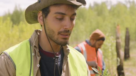 Close-up-view-of-caucasian-man-and-african-american-woman-activists-holding-small-trees-and-observing-them-to-plant-in-the-forest