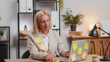 exhausted caucasian mature businesswoman with sticky notes and laptop sitting at home office desk