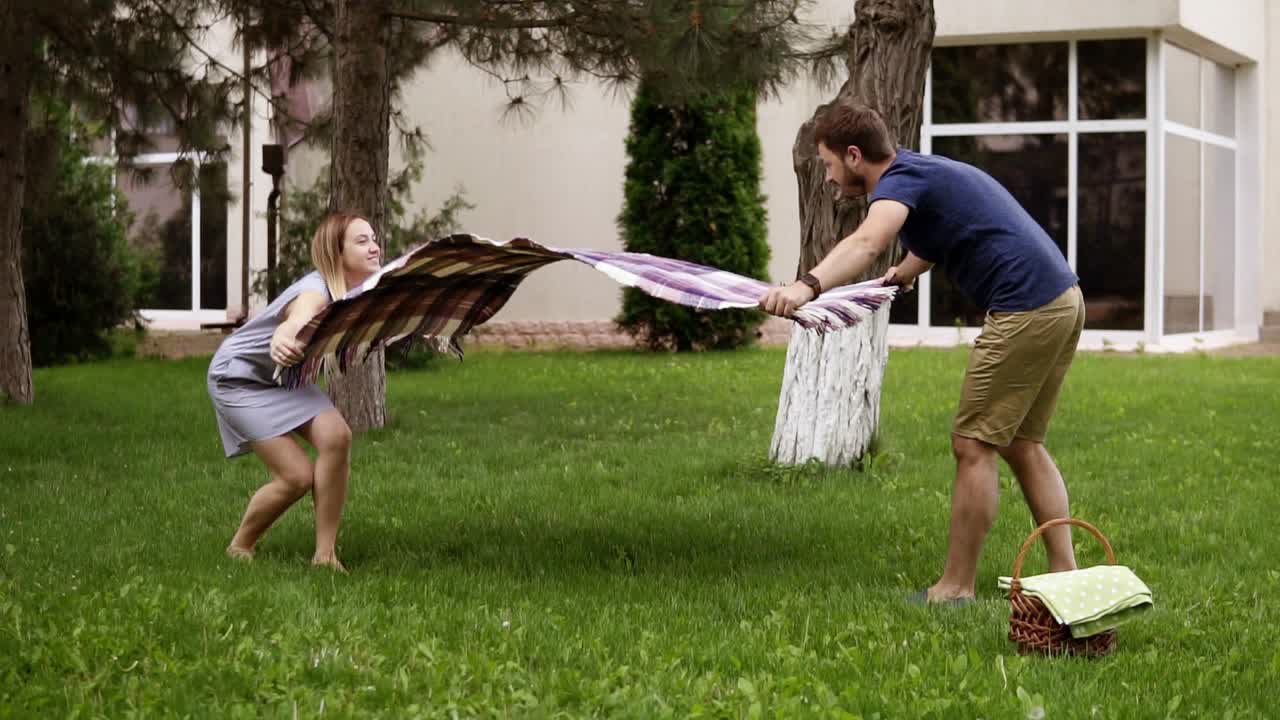 Father and mother are preparing picnic cover, laying it on grass. Lawn. Summer day. Little boy is happily running around. Slow motion
