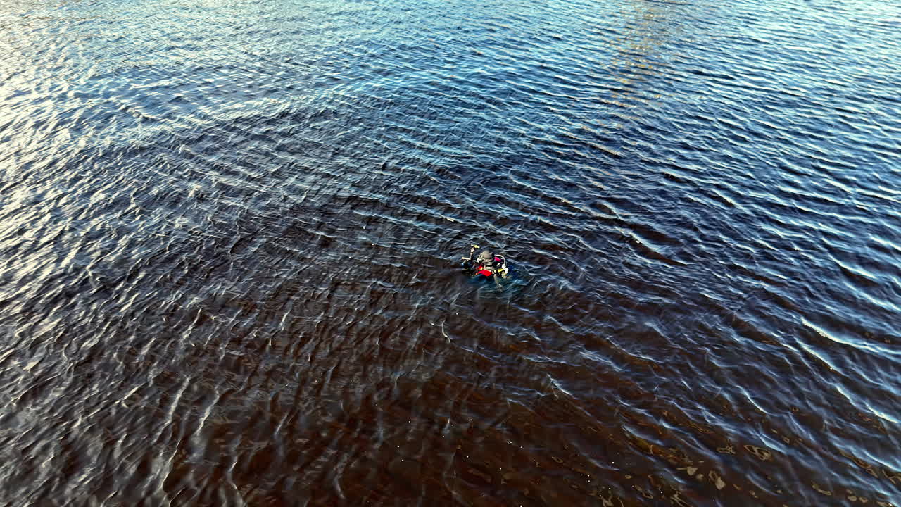 A surface diver swims across rippled open water while towing a bright marker float, captured in a wide aerial frame that emphasizes distance, situational awareness, and calm endurance