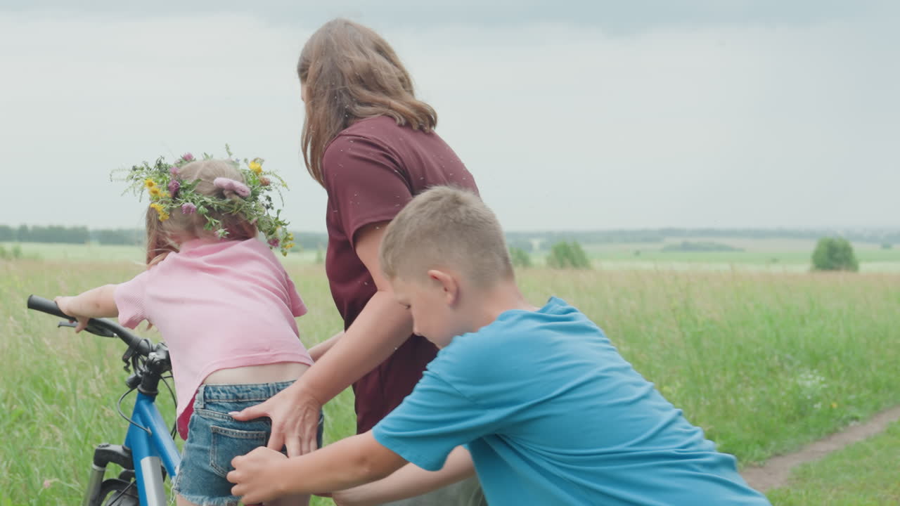 백인 엄마가 풀이 무성한 길에서 자전거를 타는 아이를 가르치고 있어요. 여자는 자전거를 잡고 있고 어린 아들은 지켜보고 있네요. 부드러운 흐린 빛, 야생화와 초원, 참을성 있는 코칭과 다정한 가족애가 느껴져요.