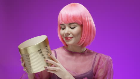 bella mujer con una peluca rosa sosteniendo una caja dorada con un regalo y sonriendo 1